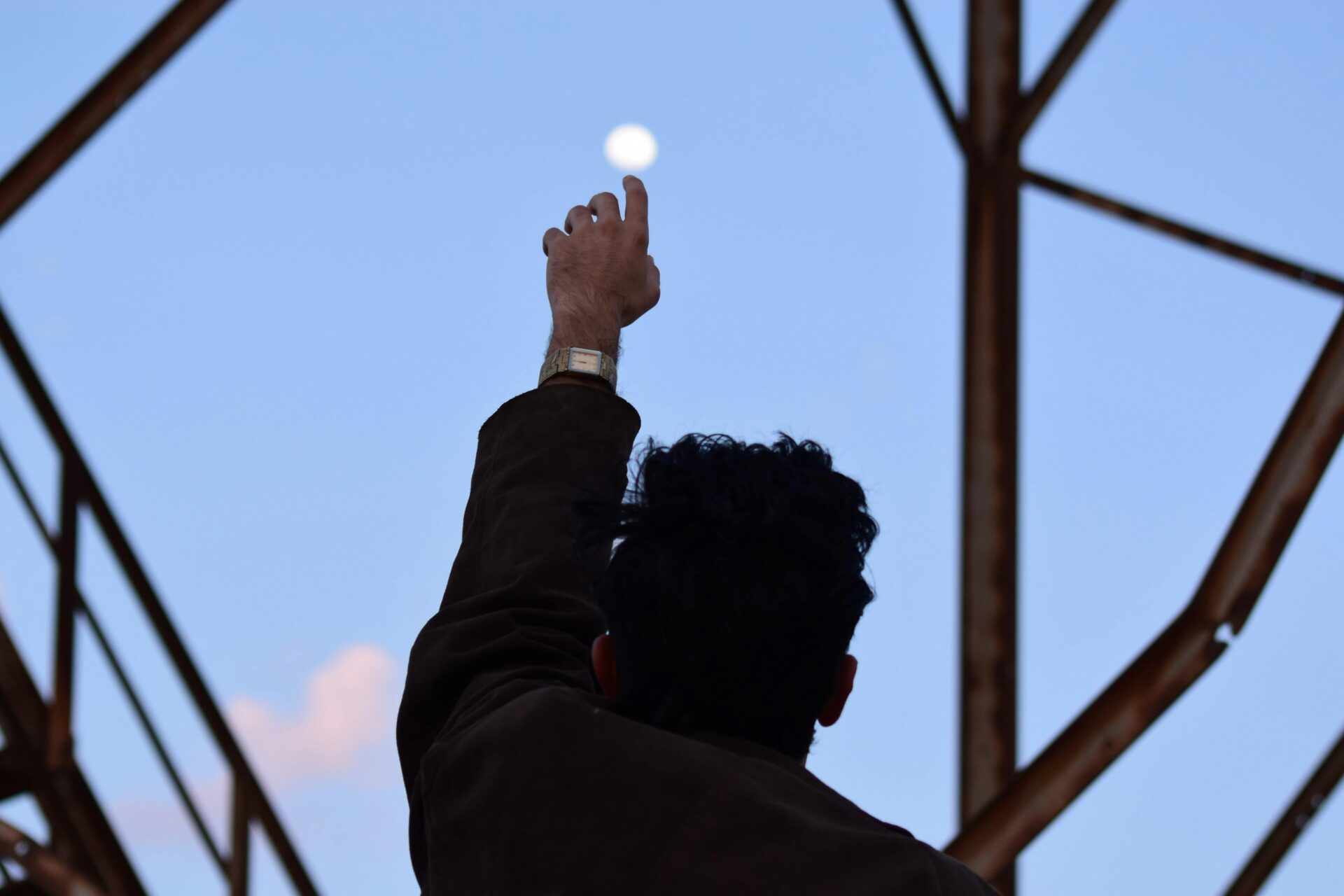 A man silhouetted against the morning sky reaches towards the moon with an outstretched hand.